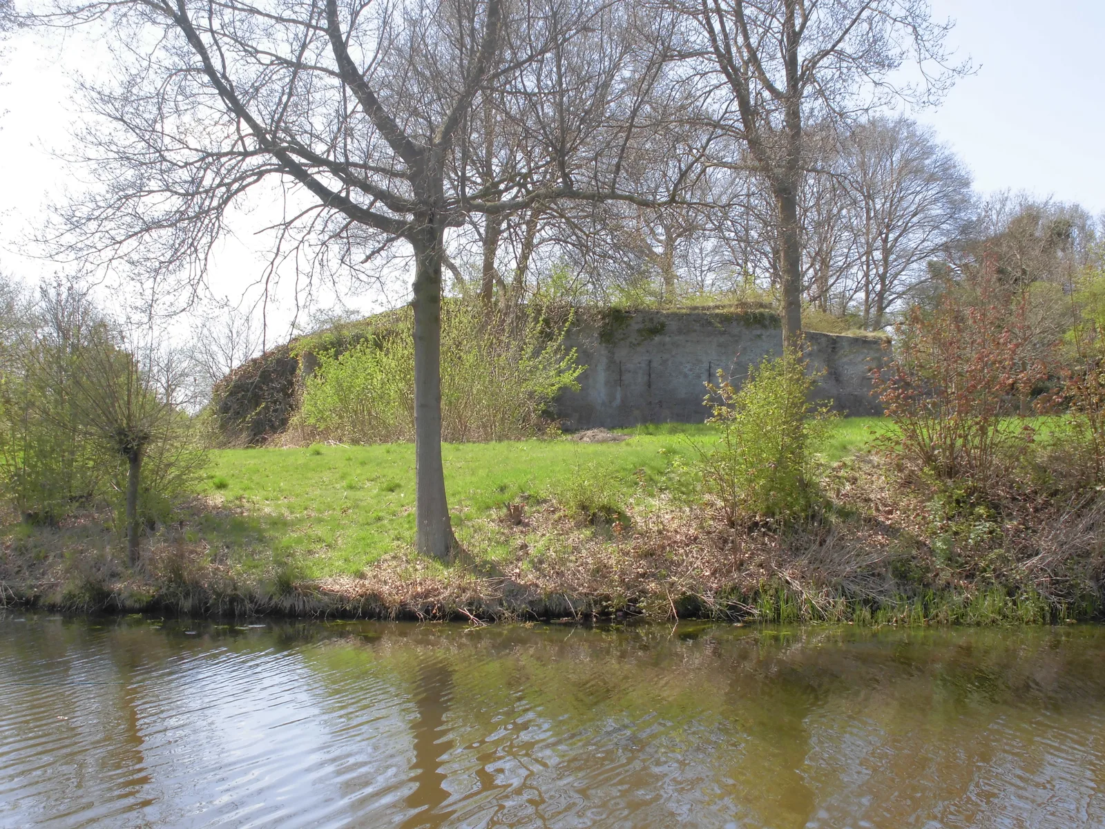 The overgrown earthwork and the mossy brick wall of Fort bij Tienhoven behind the moat